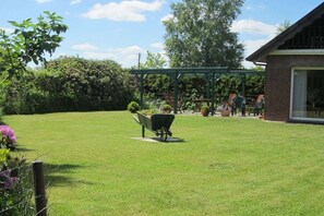 A well-maintained backyard with a green lawn, a wheelbarrow, and a brick building with a glass door. - Holiday home in Aurich with swimming pool (Aurich)