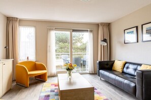 Ein modernes Wohnzimmer mit einer Couch, einem Couchtisch und einem Sessel. Ein Balkon bietet Blick auf Bäume.