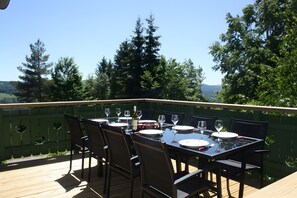 Ein Essbereich im Freien mit einem Tisch für sechs Personen, umgeben von Stühlen, auf einer Holzterrasse mit Blick auf Bäume und Hügel.