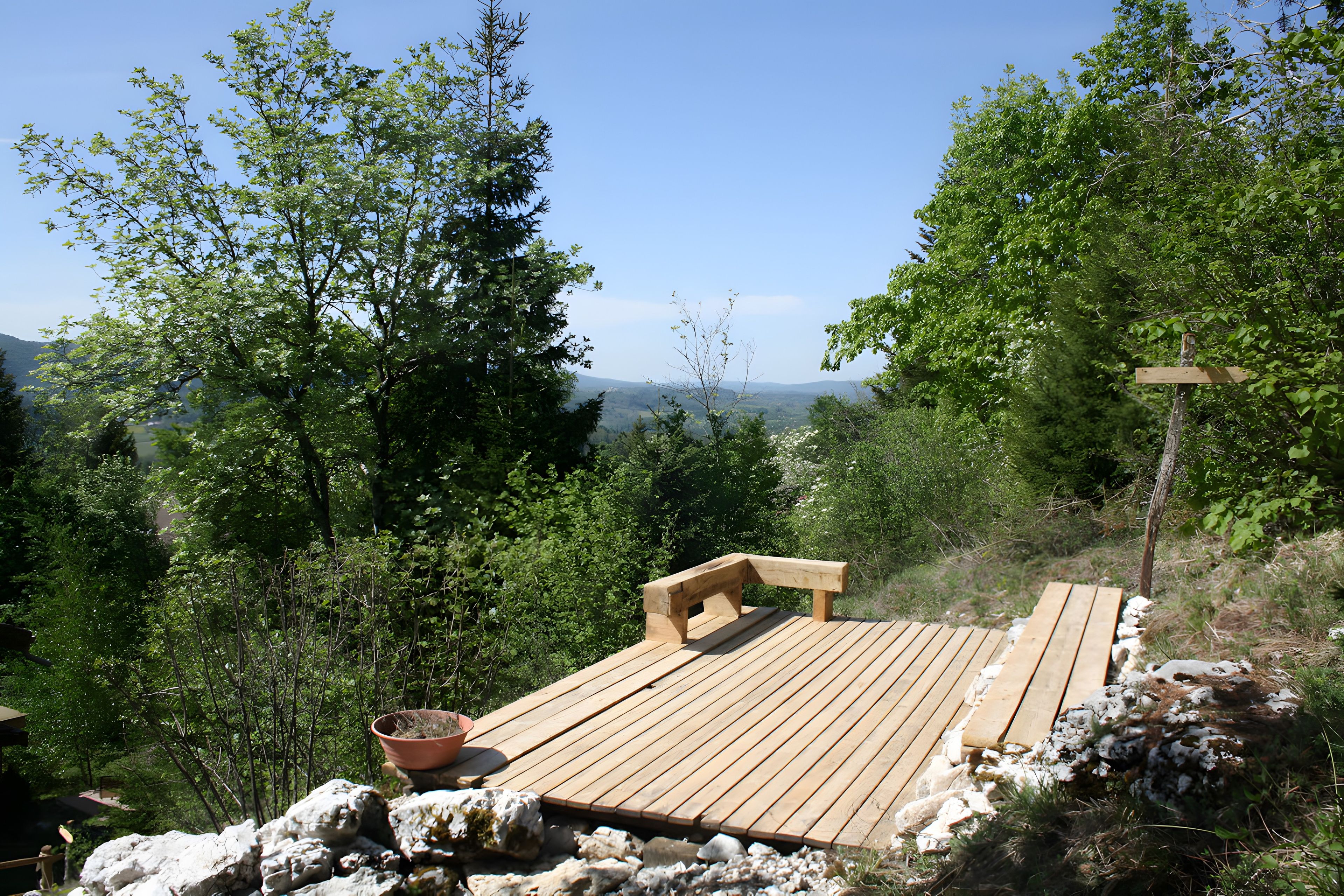 Una terraza de madera con un banco, rodeada de árboles y vistas a colinas distantes.
