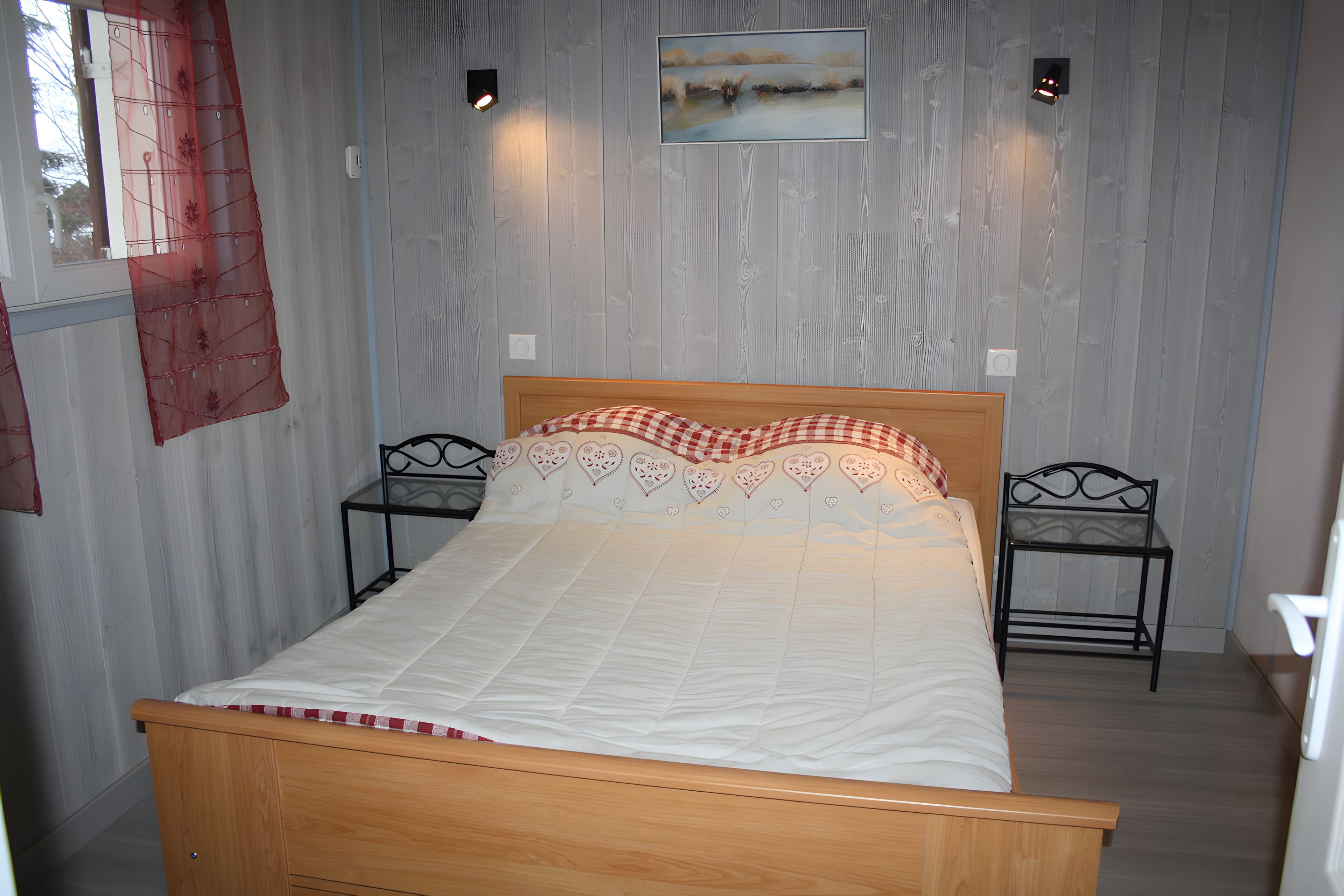 A bedroom with a wooden bed, two bedside tables, a wall-mounted painting, and a window with a curtain.