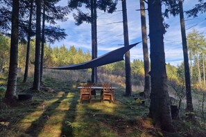 Ein Esstisch mit Stühlen in einer Waldlandschaft, umgeben von hohen Bäumen und einem hängenden Lampenschirm.