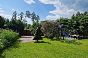 Um jardim bem cuidado com diversas árvores, um pequeno pinheiro de Natal e um parquinho com um escorrega.