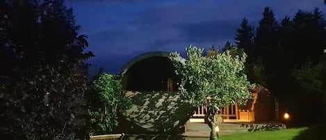 A nighttime view of a property with a covered outdoor seating area, a wooden building, and a tree in the foreground.
