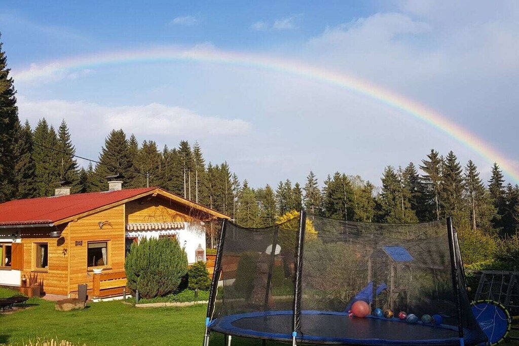 Une cabane en bois avec un toit rouge, un trampoline doté d’un filet de sécurité et un arc-en-ciel en arrière-plan.