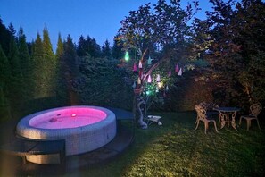 Ofurô circular com luz rosa, cercado por gramado, cadeiras e árvore com luzes penduradas.