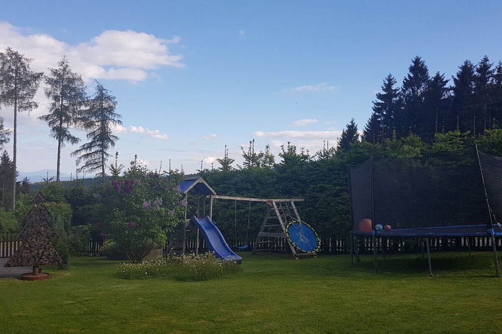 Un parque infantil con tobogán, columpios y trampolín en un césped arbolado.