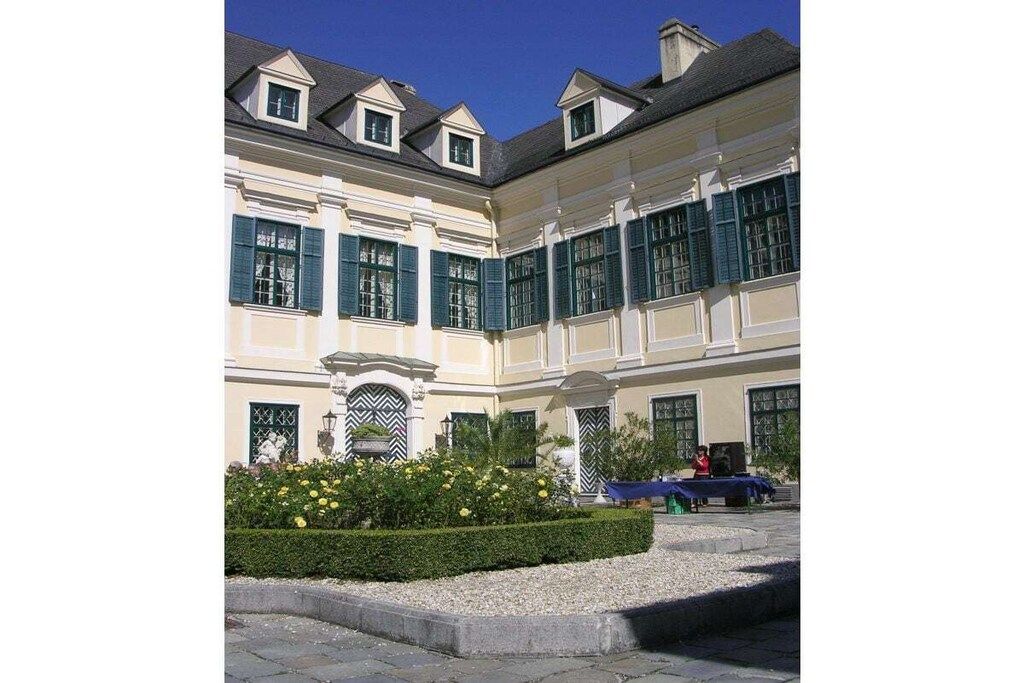 Un bâtiment de style classique, doté d’une architecture symétrique, de nombreuses fenêtres avec volets et d’un jardin bien entretenu agrémenté de fleurs jaunes.