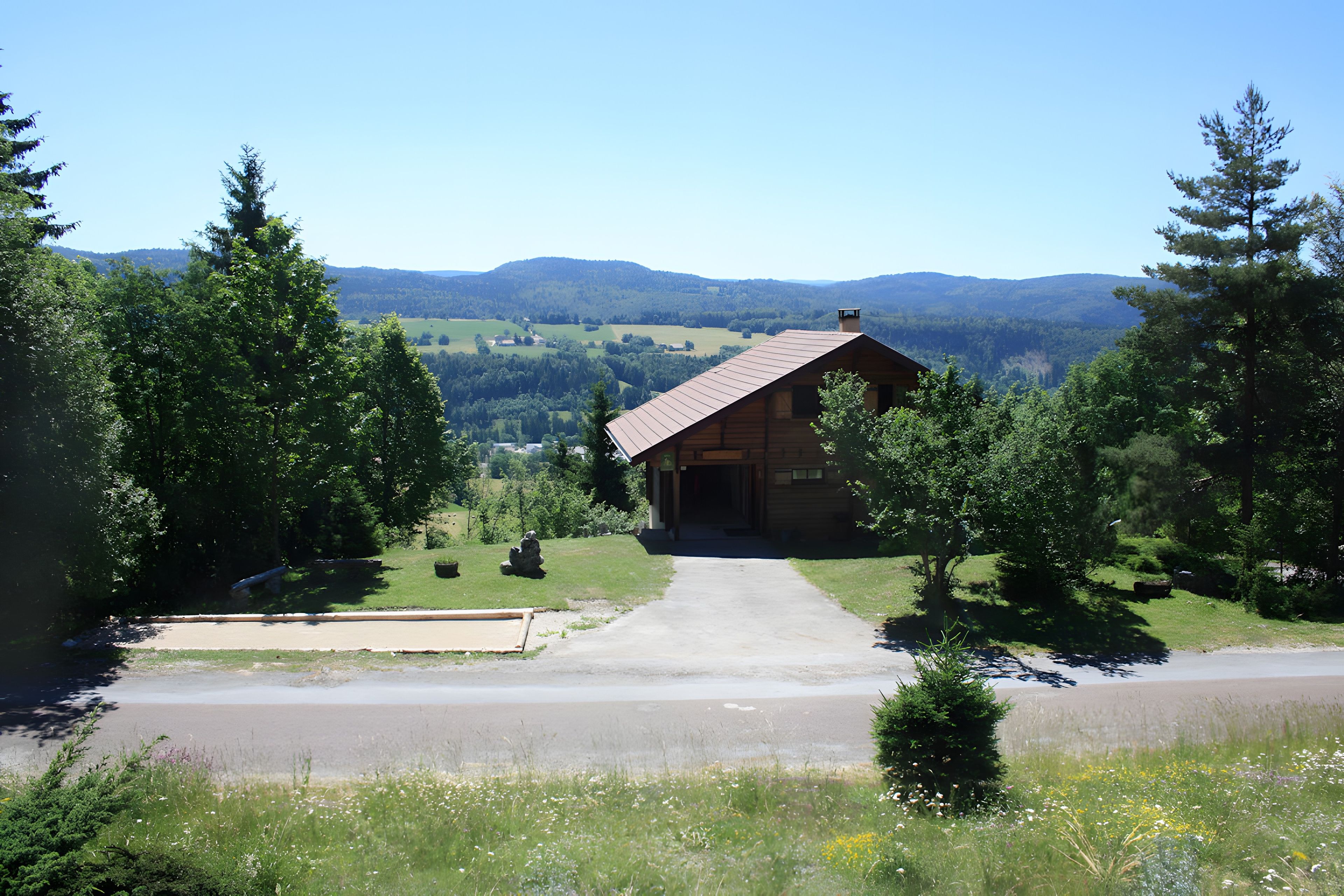 A wooden cabin with a metal roof, surrounded by lush greenery and a clear view of distant hills.