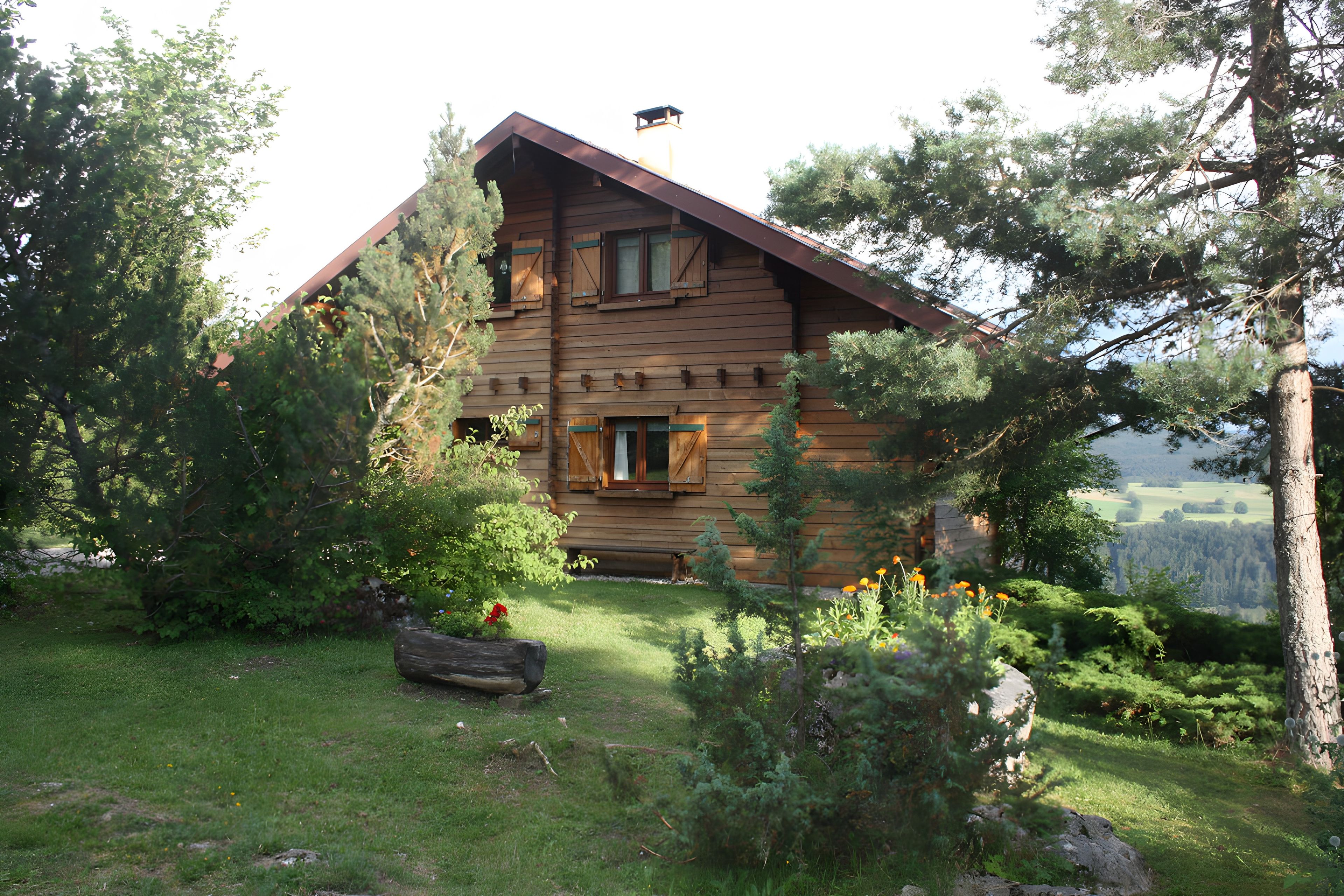 Eine Blockhütte mit geneigtem Dach, umgeben von einem gepflegten Garten und Bäumen.