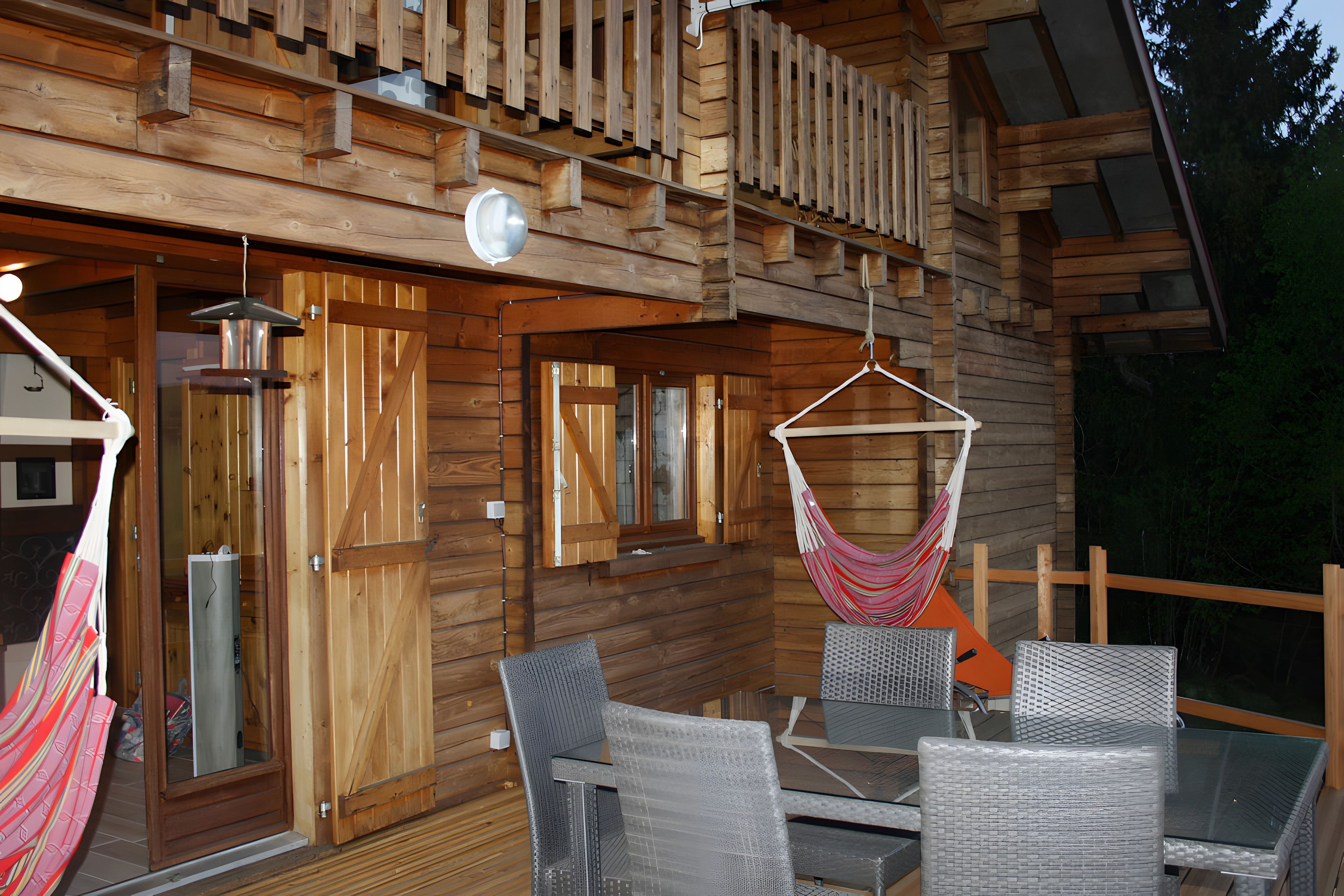 A wooden cabin with a balcony, a hanging hammock, and a glass table with chairs.