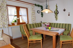 Ein Essbereich mit einem Holztisch und grünen, gepolsterten Stühlen, einem Fenster mit Vorhängen, einer Pendelleuchte und einer Wanduhr.