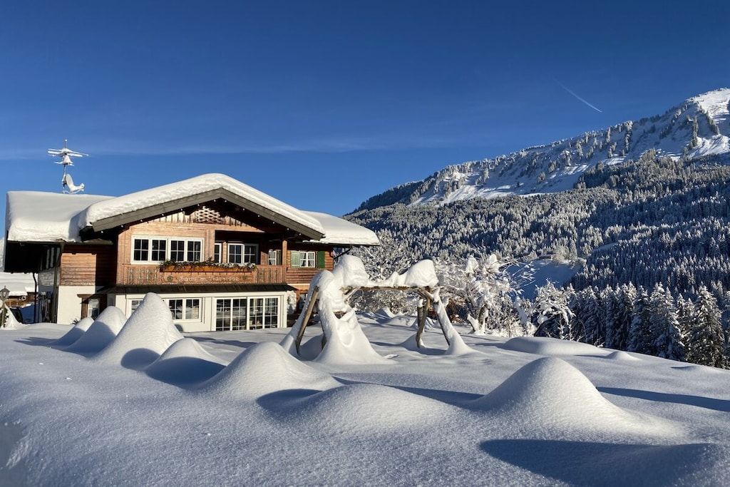 A wooden chalet with a sloped roof, surrounded by snow-covered trees and a mountain backdrop.
