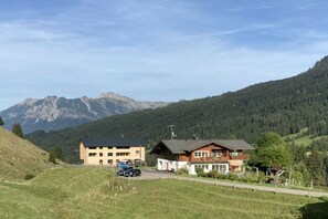 A two-story building with a sloped roof, a parking area, and a view of a mountain range. - Fellhorn in the House of Narcissus (Riezlern)