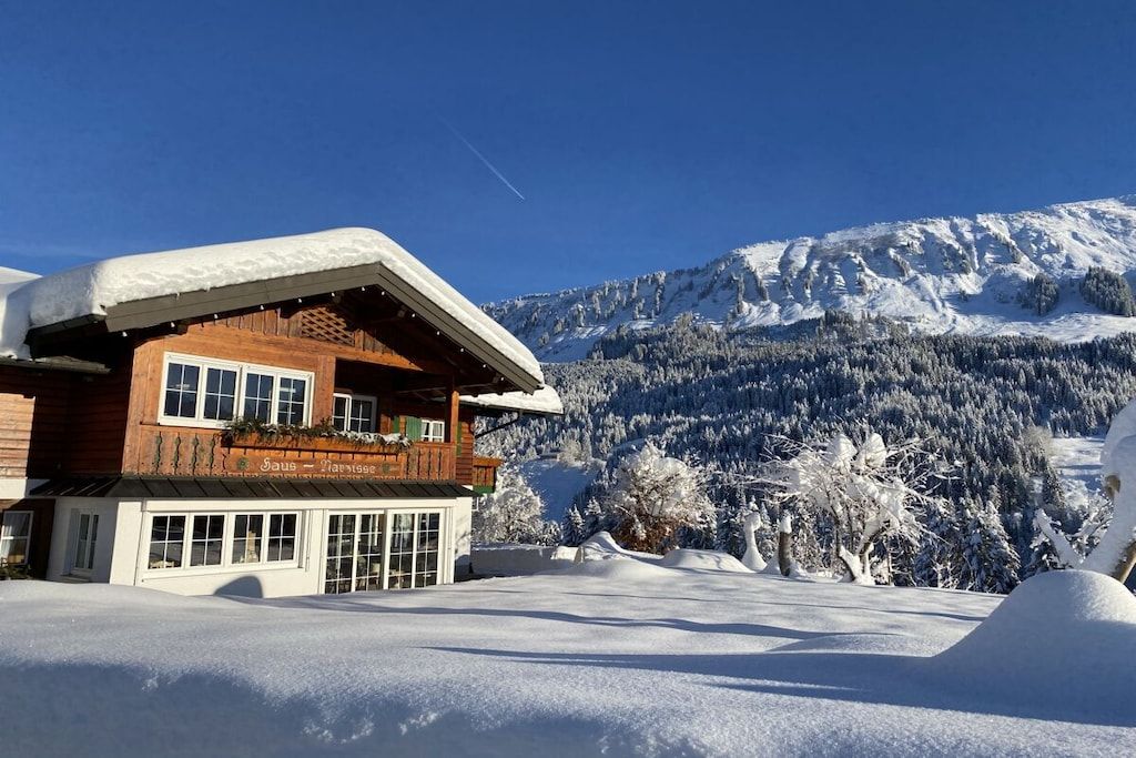 Un chalet en bois doté d’un balcon, entouré de montagnes et d’arbres couverts de neige.