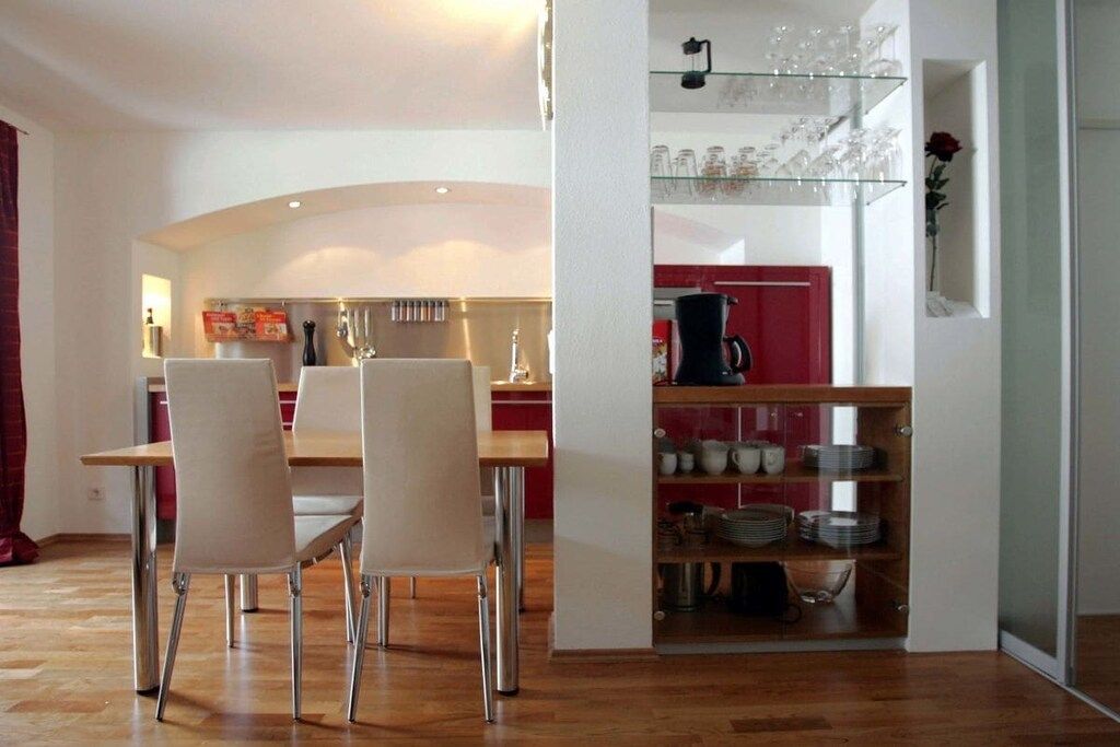 A dining area with a wooden table and chairs, a kitchen area with red cabinets, and a glass display shelf with kitchenware.