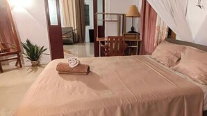Una cama bien hecha con una toalla doblada, una silla de madera, una planta pequeña en maceta y un escritorio con una lámpara.