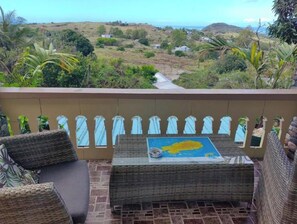 A balcony with wicker furniture, a table with a map, and a view of a hilly landscape. - Henz & Sia Tourist Residence – Your Island Home in Rodrigues 🌴 (Piments Reposoir Rodrigues)