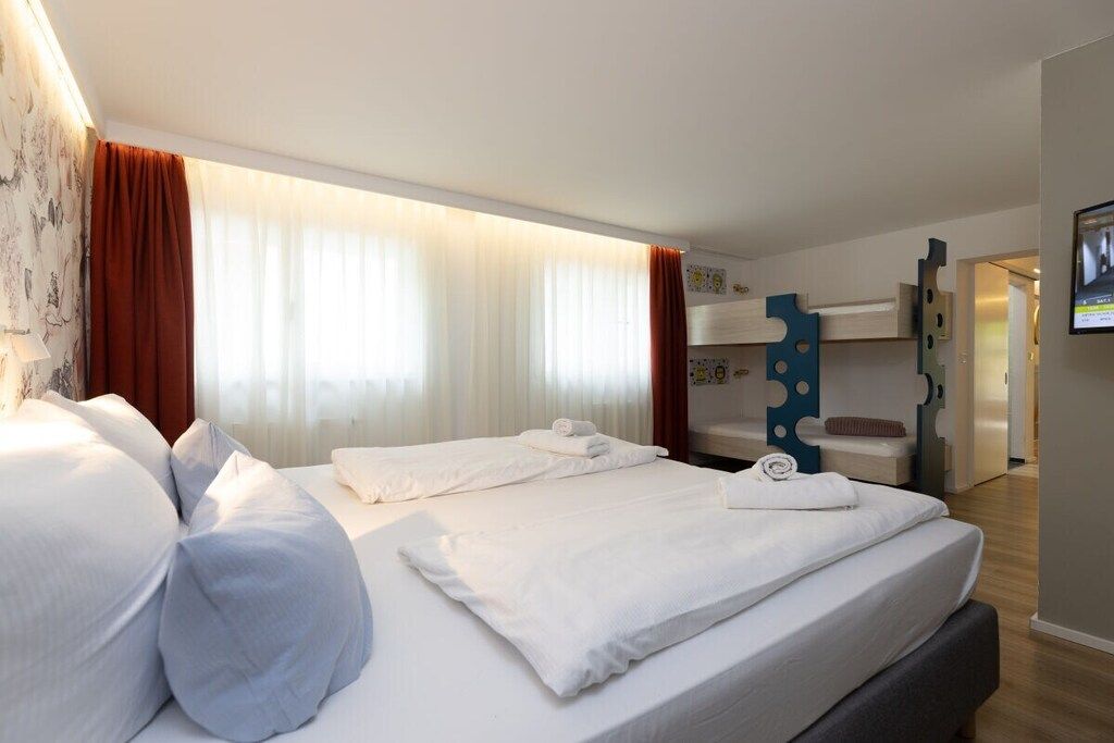 A hotel room with two beds, a bunk bed, a bathroom with a mirror, and a wall-mounted TV.