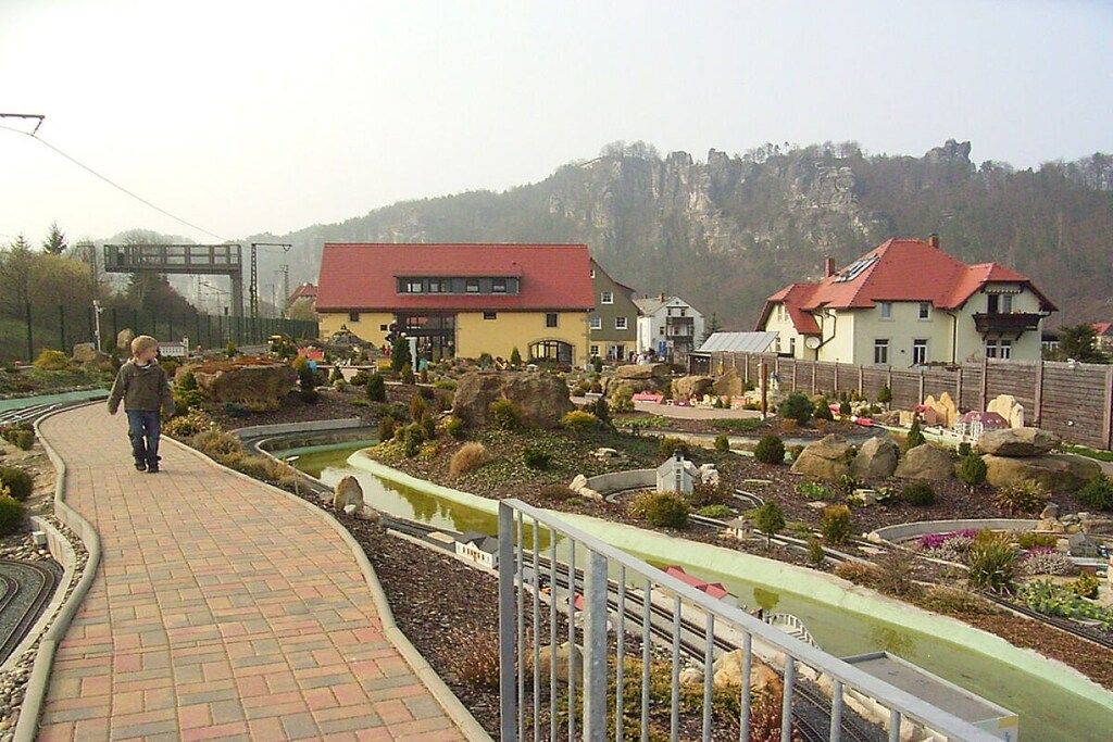 A paved pathway with a small train track, surrounded by landscaped gardens and buildings with red roofs.