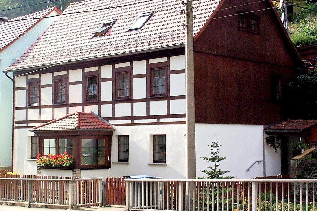 Ein traditionelles Fachwerkhäuschen mit geneigtem Dach, einer kleinen Veranda mit einem Fenster und einem Holzzaun davor.