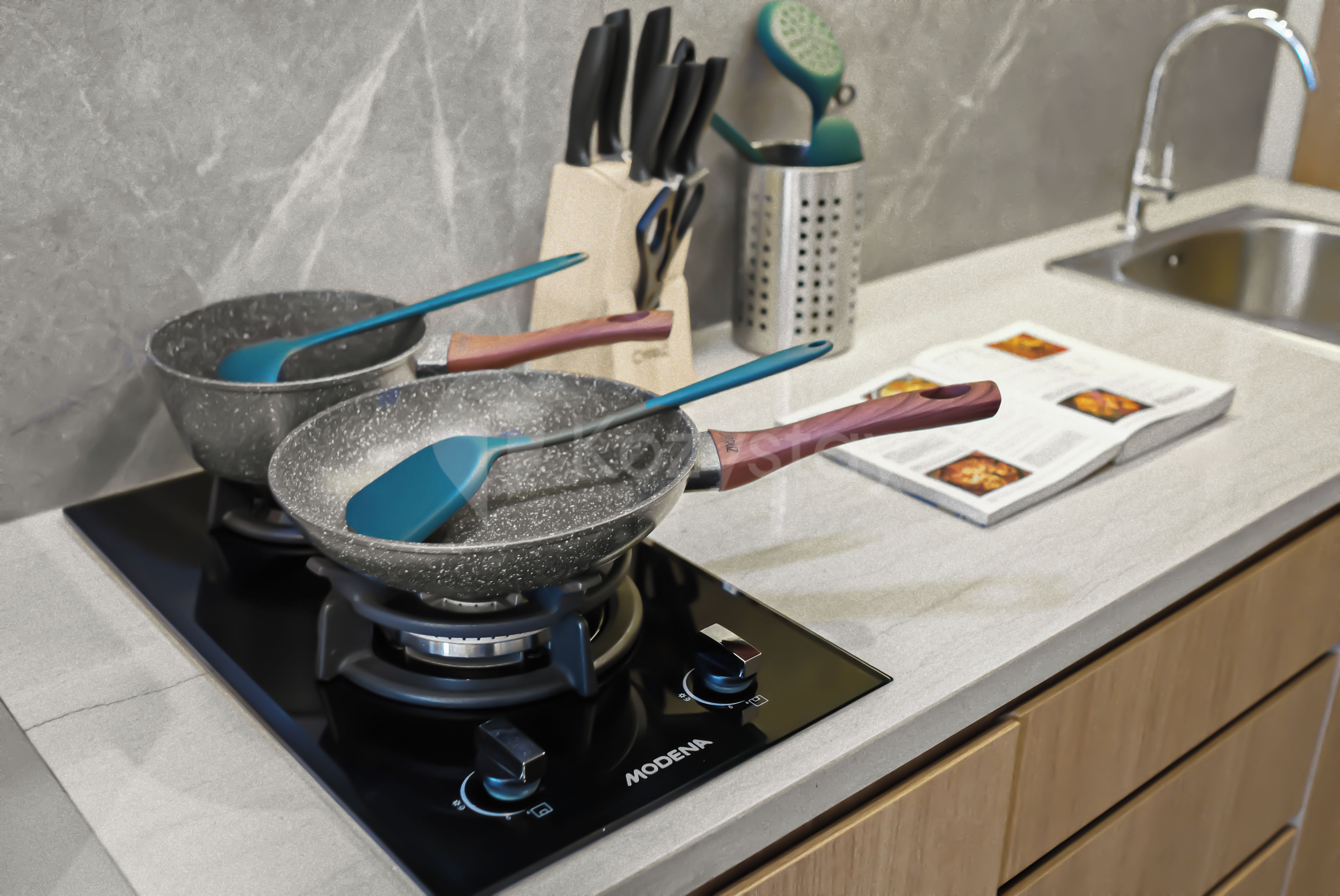 Una cucina con un fornello, due padelle, una spatola e un libro di cucina sul bancone.