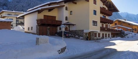 A snow-covered building with a sloped roof, a balcony, and a mountain backdrop.