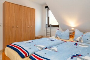 A bedroom with a wooden wardrobe, a bed with a sailboat pattern bedspread, and a window with a view of the outside. - House Ahrens Apartment 4 (Zingst (Ostseeheilbad))