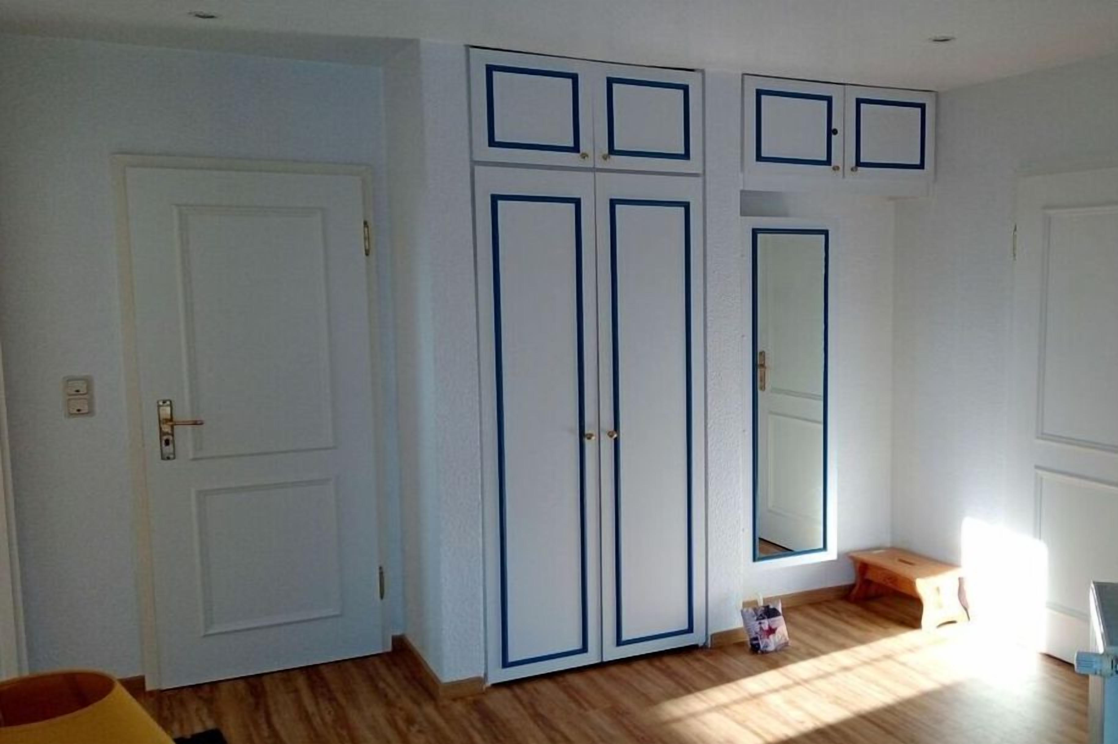 Ein Zimmer mit einer weißen Tür, einem Schrank mit blauer Zierleiste, einem Spiegel und einem kleinen Holzschemel.