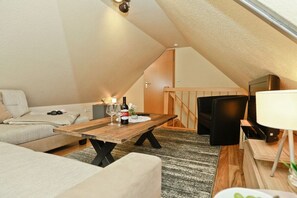 Et hyggeligt loftrum med et træspisebord, en sofa, et fjernsyn og en trappe op til et hvileområde.