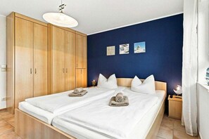 Ein Doppelbett mit weißen Bettwäsche und zwei Handtüchern, ein Holzschrank, ein Nachttisch mit Lampe sowie eine Wand mit drei gerahmten Bildern.
