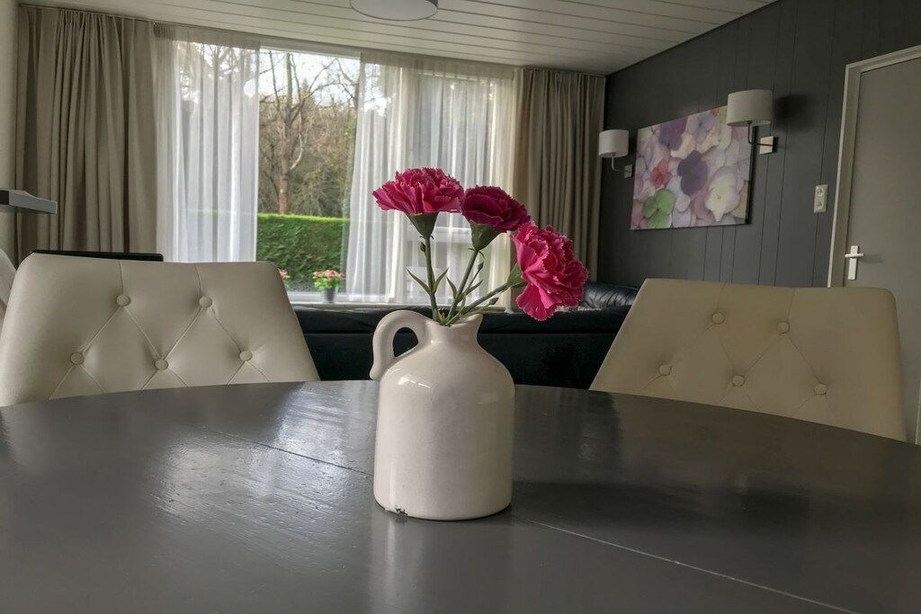 Et spisebord med en hvid kande og lyserøde blomster, en polstret bænk og et vindue med gardiner.