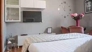 Ein Hotelzimmer mit Bett, Schreibtisch, Fernseher, einer Uhr und einer Blumenvase.