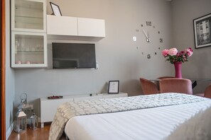 A hotel room with a bed, a desk, a television, a clock, and a vase with flowers. - Vrbo Property (Roma)