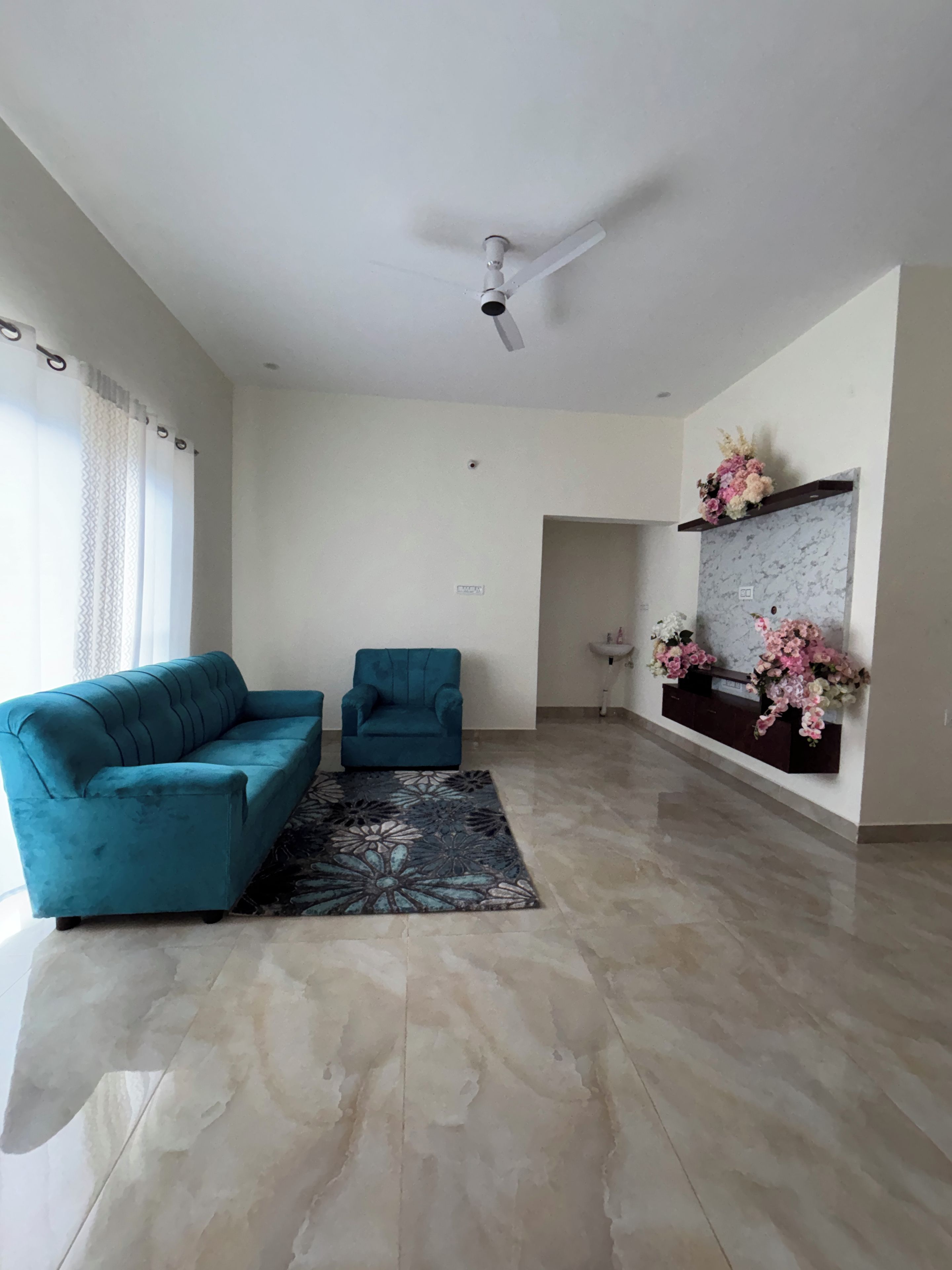 A spacious living room with a teal sofa, a smaller armchair, a patterned rug, a ceiling fan, and a wall-mounted shelf with flowers.