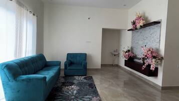 A spacious living room with a teal sofa, a smaller armchair, a patterned rug, a ceiling fan, and a wall-mounted shelf with flowers.