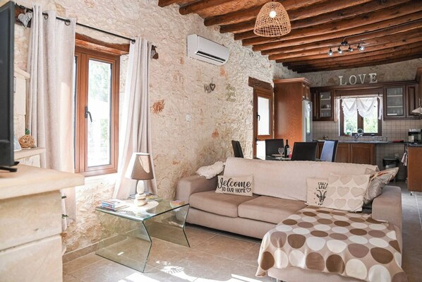 A cozy living room with a sofa, a glass coffee table, and a television. There is a kitchen area with wooden cabinets and a window.