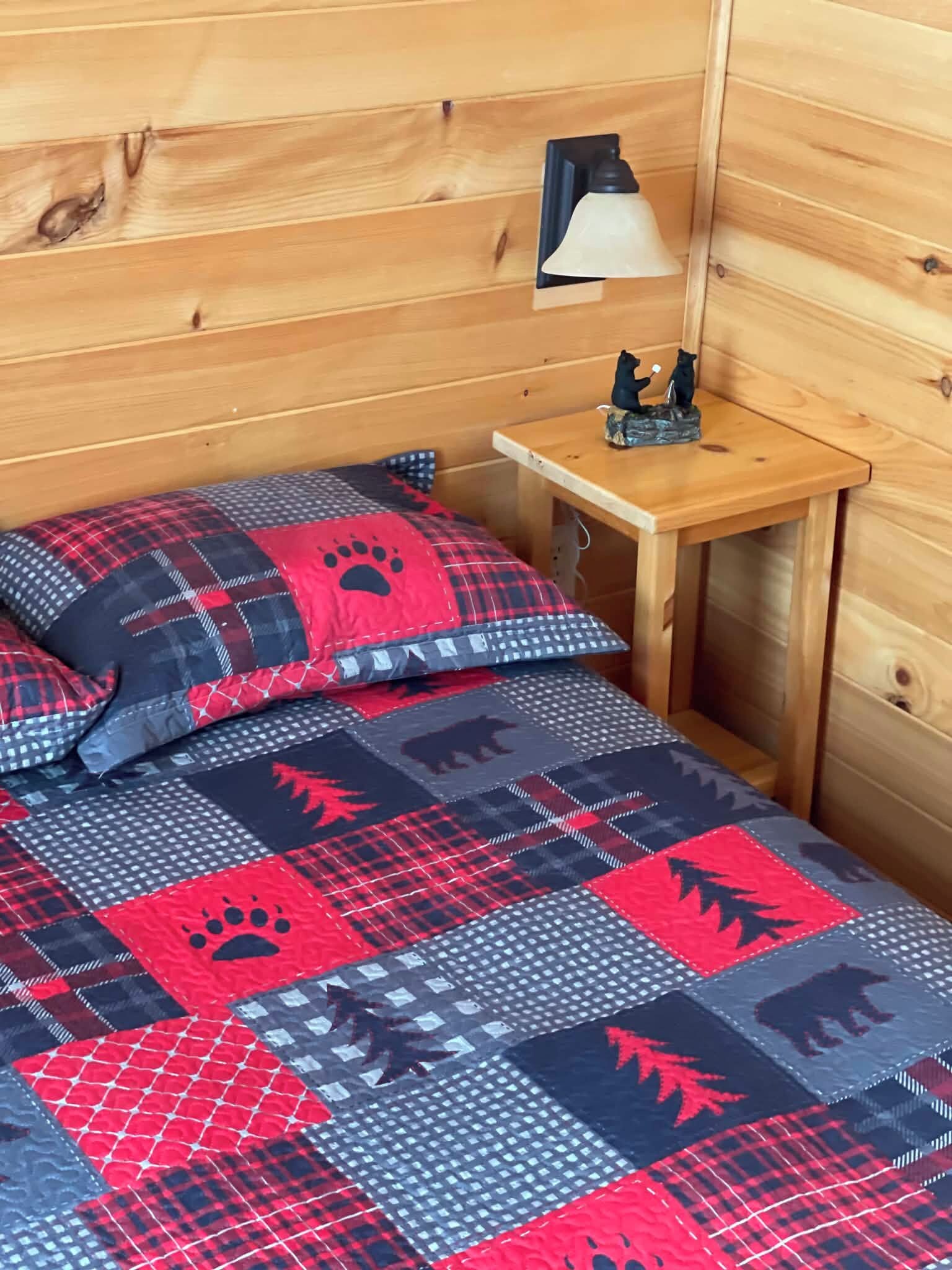 L’intérieur d’un chalet en bois comprend un lit recouvert d’une courtepointe rouge et bleue ornée d’empreintes de pattes et de motifs de feuilles, une petite table de chevet avec une lampe et une figurine d’ours.
