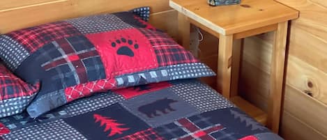 A wooden cabin interior with a bed featuring a red and blue quilt with paw prints and leaf patterns, a small bedside table with a lamp, and a bear figurine.