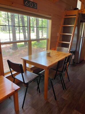 Interior de madera con una mesa, sillas, un refrigerador y una ventana con vista a árboles.