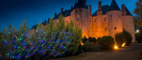 A castle with a moat, illuminated by warm lights, and surrounded by trees decorated with colorful lights.