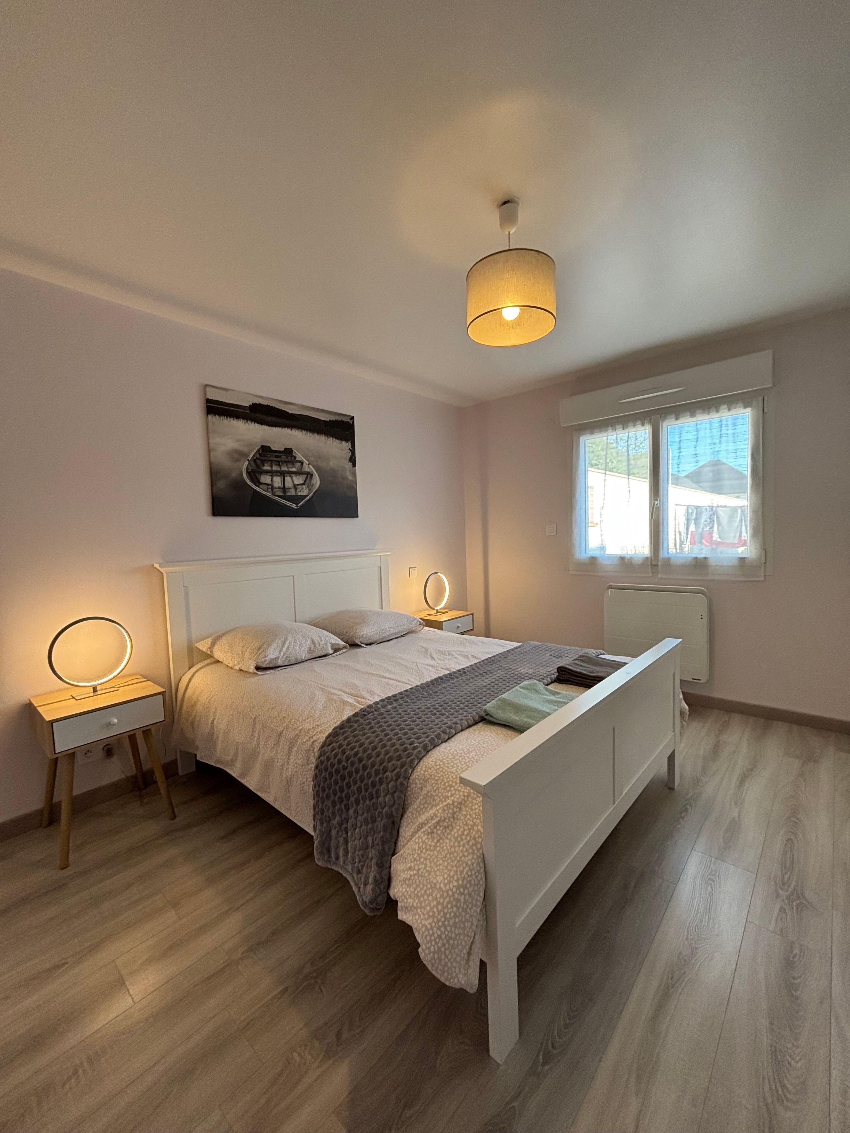 Een slaapkamer met een bed, nachtkastjes, een hanglamp, een raam met gordijnen en een aan de muur hangend schilderij.