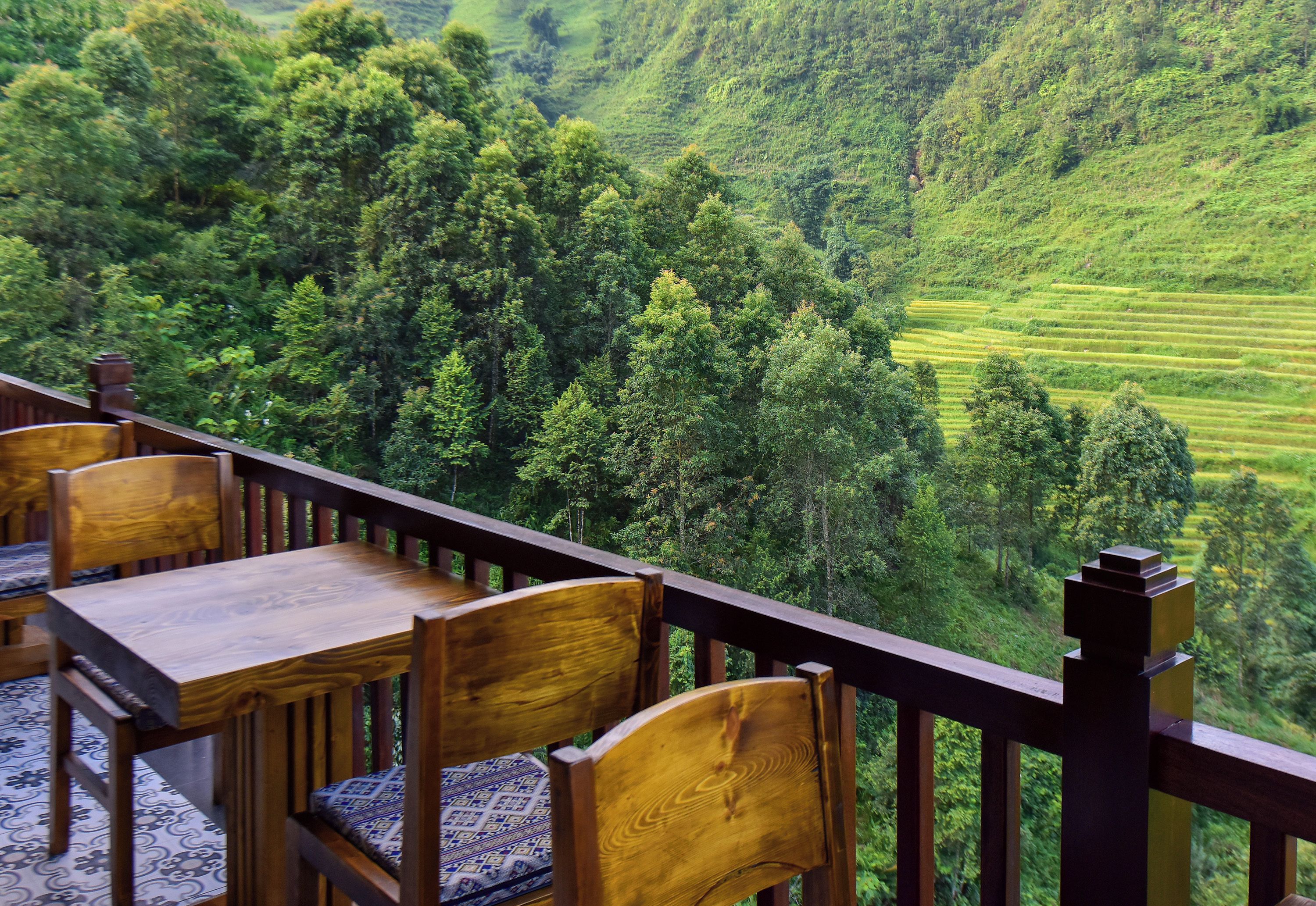 A wooden deck with a table and chairs, overlooking a lush green valley with terraced fields.