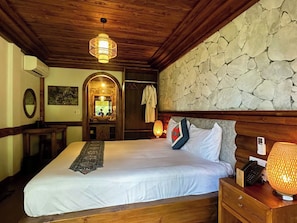 Habitación de hotel con una cama grande, cabecera de madera, pared de piedra y un pequeño escritorio con lámpara.