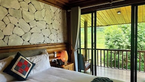 A hotel room with a stone wall, a bed with a quilted pillow, a nightstand with a lamp, and a view of greenery through a balcony. - MAISON DE LAO CHAI (Sa Pa)