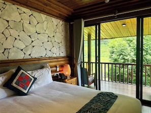 Habitación de hotel con pared de piedra, cama con almohada acolchada, mesita de noche con lámpara y vistas a la vegetación desde un balcón.