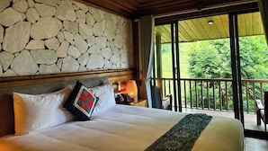 A bedroom with a stone wall, a bed with pillows, a nightstand with a lamp, and a balcony with a view of greenery. - MAISON DE LAO CHAI (Sa Pa)