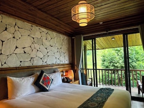 Un dormitorio con pared de piedra, una cama con almohadas, una mesita de noche con lámpara y un balcón con vista a follaje.