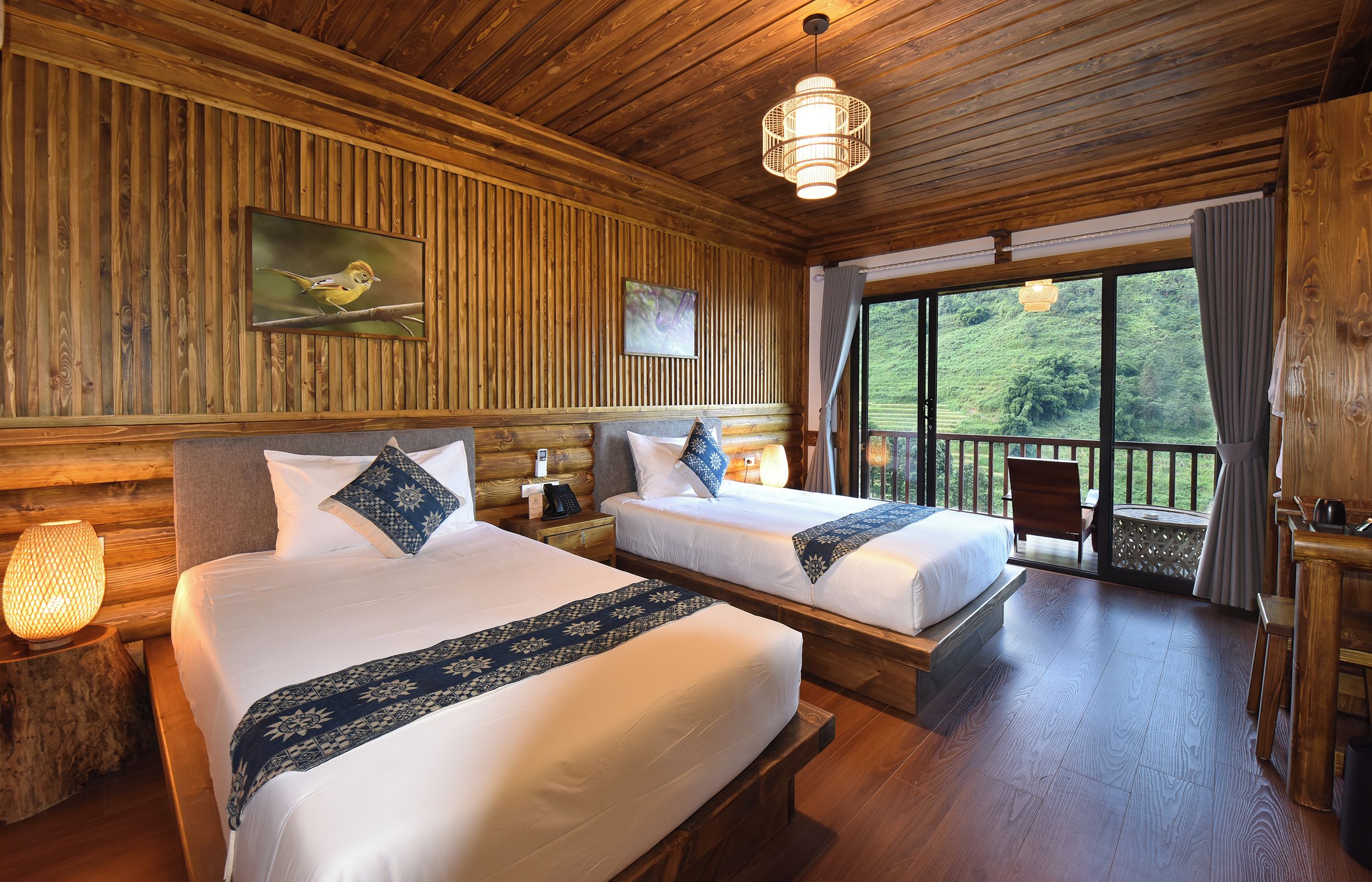 A wooden cabin room with two beds, a wooden ceiling, a large window, and a balcony.