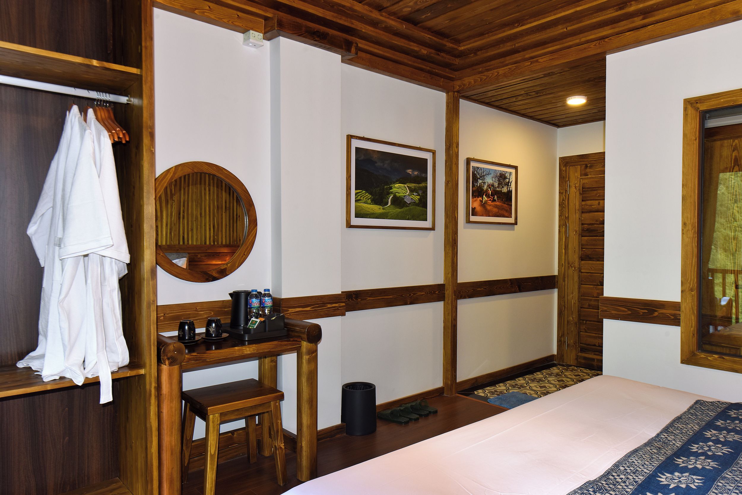 A room with a bed, a wooden desk with a mirror, a coat rack, and two framed pictures on the wall.